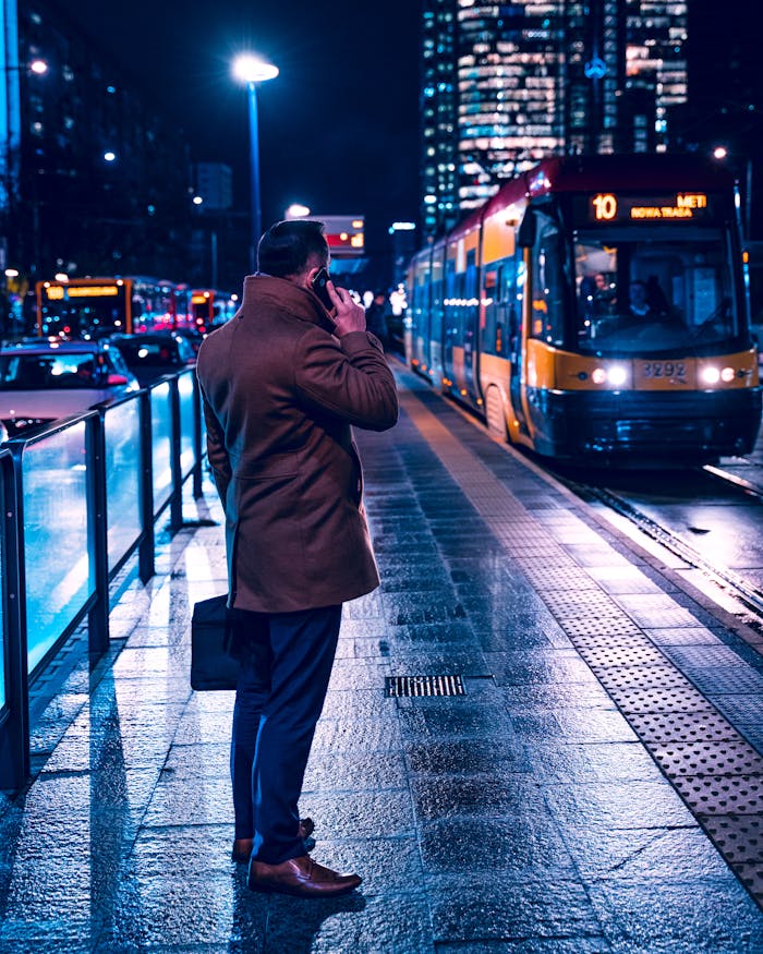 A professional man in a coat makes a phone call at a tram stop in Warsaw at night.
